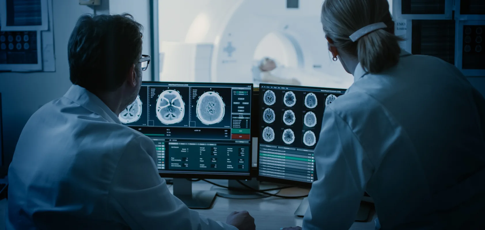 Two medical radiation technologists reviewing CT brain scans on monitors beside a scanner suite